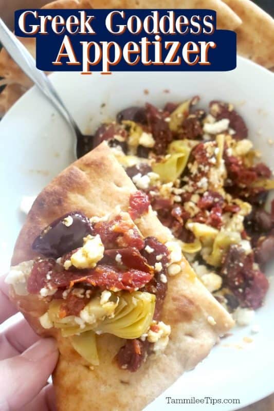 Greek goddess appetizer over a hand holding pita covered in kalamata olives, sundried olives, and feta