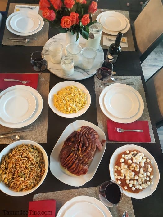 Dining room table with candles, roses, white plates, ham, green bean casserole, corn, and sweet potato casserole