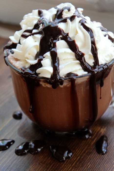 Hot chocolate in a glass mug garnished with a heaping pile of whipped cream and chocolate syrup 