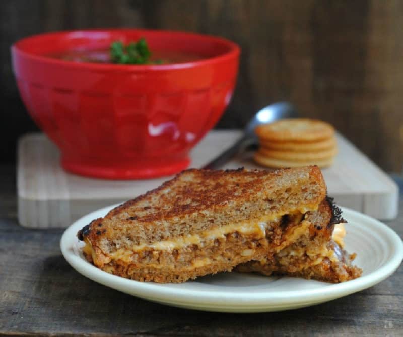 Grilled cheese sloppy joe on a white plate next to a bowl of tomato soup and crackers