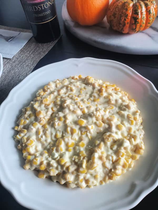 Crockpot cream corn in a white serving bowl with 2 pumpkins and a bottle of wine behind it. 
