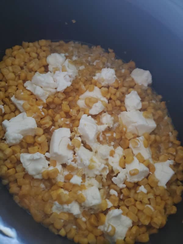 crockpot cream corn in a black slow cooker, chunks of cream cheese spread over the corn kernels before cooking. 