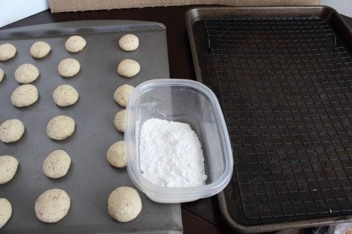Snowball cookies on a baking sheet next to a bowl of powdered sugar and a drying rack