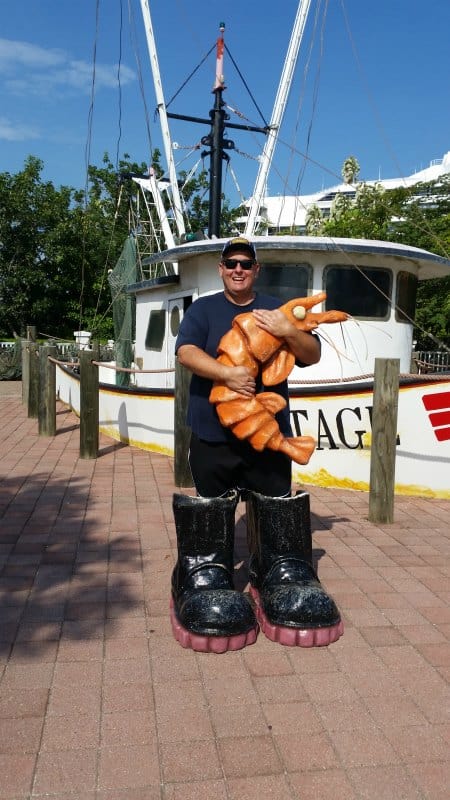 John is big fake boots holding a shrimp with a shrimp boat behind him