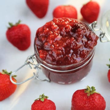 slow cooker strawberry jam in a glass storage container surrounded by strawberries