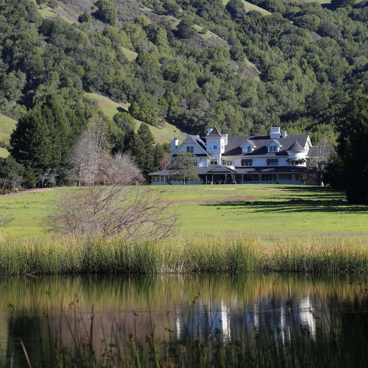Large white house in the background by rolling green hills with a lake in the front with a reflection of the house.
