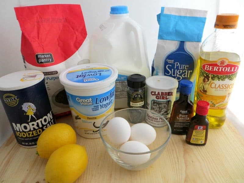 flour, milk, salt, yogurt, sugar, oil, baking powder, lemons, eggs, poppy seeds, vanilla, and almond extract on a cutting board. 
