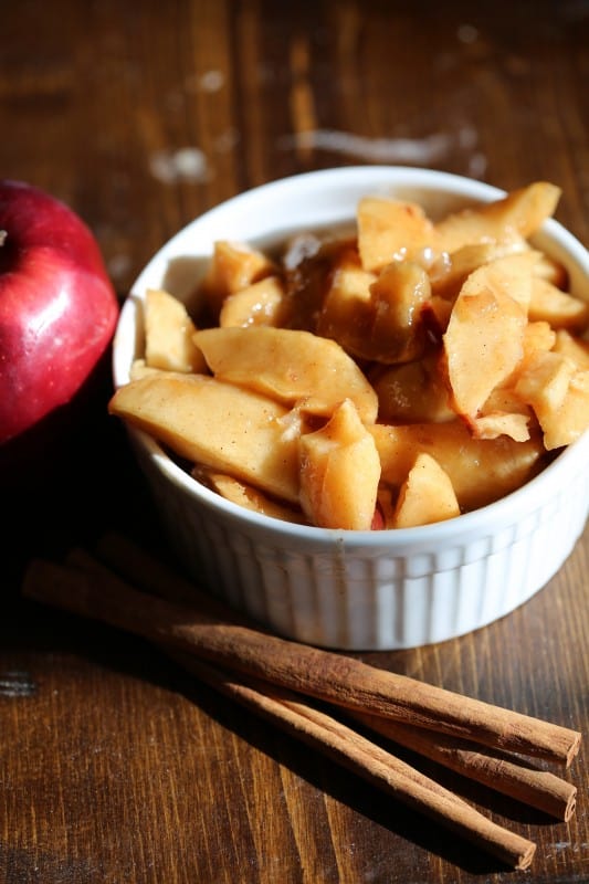 Fried apples in a white bowl next to cinnamon sticks and a red apple 