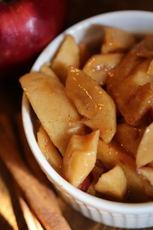 Slow cooker fried apples in a white bowl next to cinnamon sticks and a red apple 