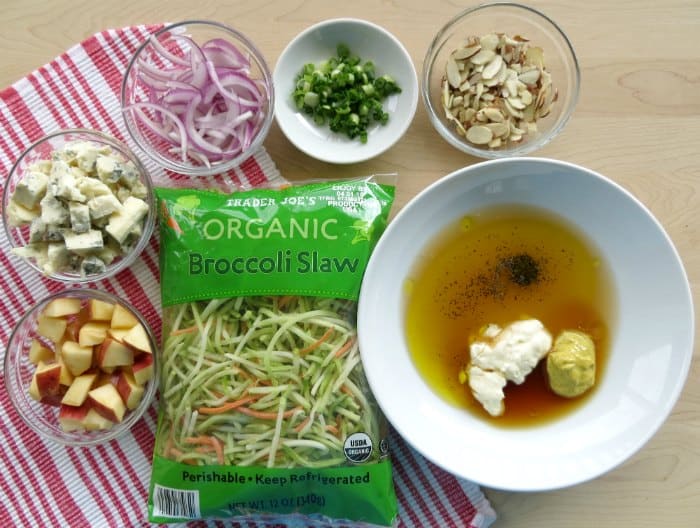 Broccoli Slaw ingredients on a wood cutting board, apples, blue cheese, red onions, slivered almonds, and more