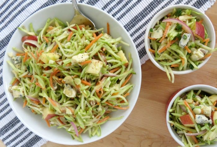 broccoli slaw in a white serving bowl with a silver spoon next to two smaller bowls on a striped napkin