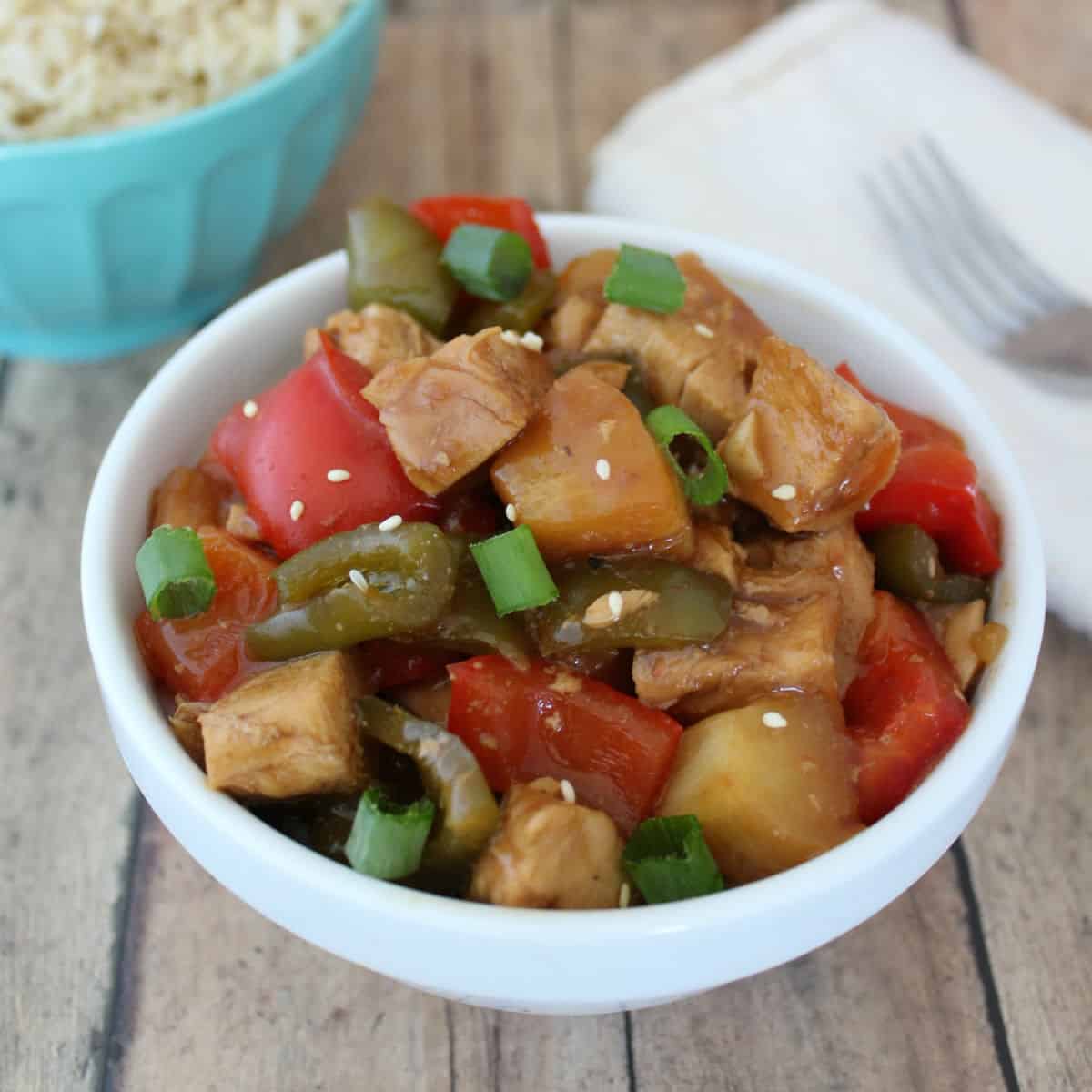 Crockpot Sweet & Sour Chicken in a white bowl next to a napkin and a fork and a bowl of rice