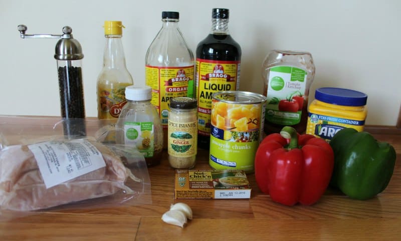 Sweet and Sour Chicken ingredients on a wood counter including pineapple, peppers, chicken breast, garlic, cornstarch, pepper,, honey, ginger, and more