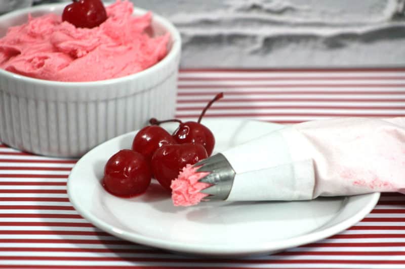 cherry frosting in a white bowl next to a plate with maraschino cherries and an icing dispenser. 