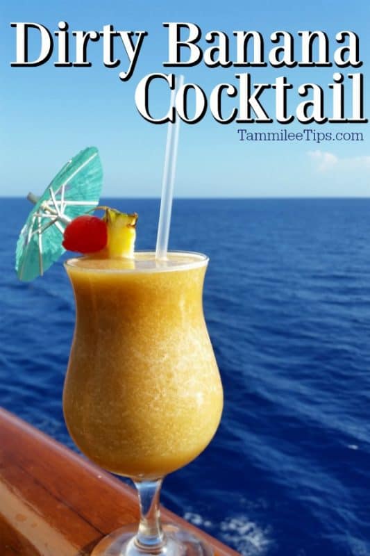 Dirty Banana Cocktail over a hurricane glass with a yellow drink, umbrella, and cherry pineapple garnish sitting on a rail with the ocean in the background. 