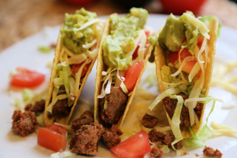 Three ground beef tacos with guacamole, tomatoes, and cheese in taco shells