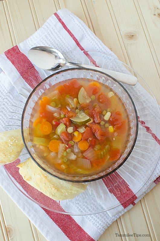 vegetable soup in a glass bowl sitting on a glass plate next to a silver spoon and cloth napkin