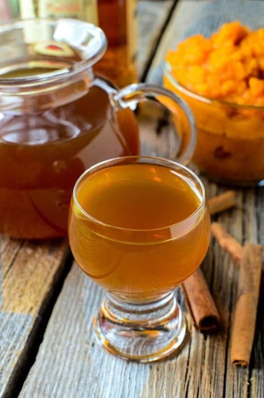 Small glass of Pumpkin Spice Liqueur on a wood background with cinnamon sticks, a bowl of pumpkin puree, and pitcher