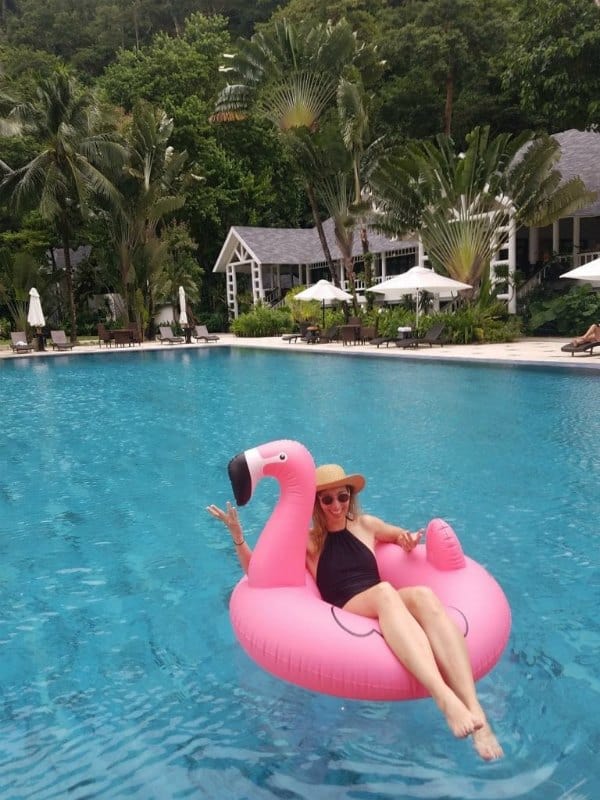 Woman riding a pink flamingo in a large pool with palm trees in the background
