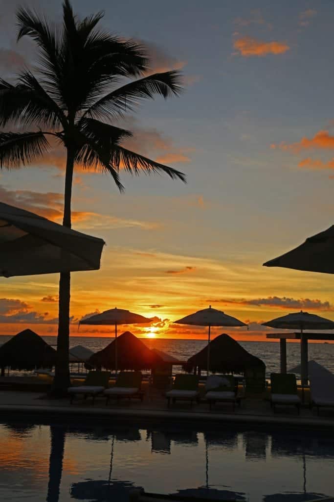 Sunset over palm trees, a pool, and umbrellas 