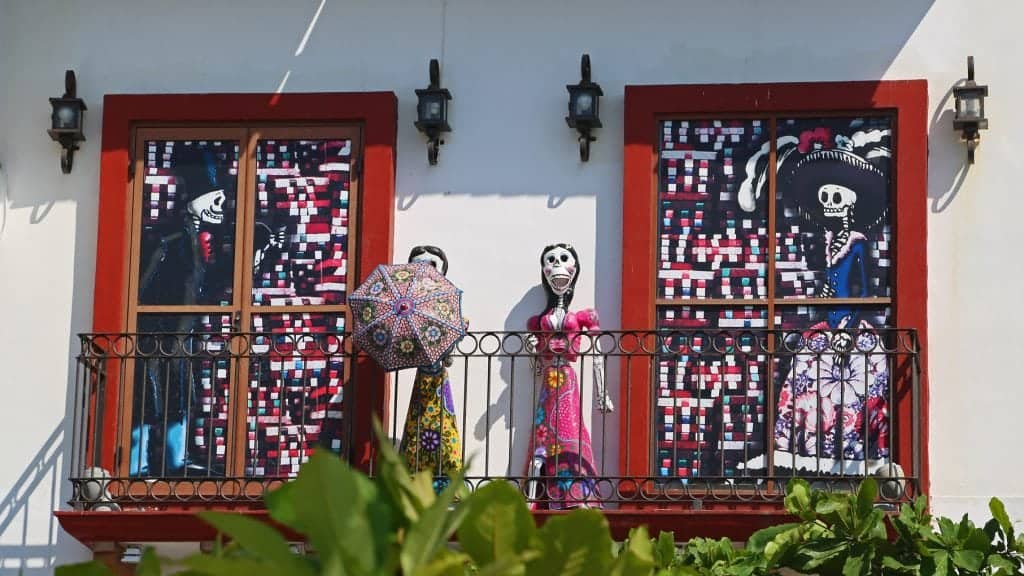 Day of the dead statues on a balcony next to two windows with paintings