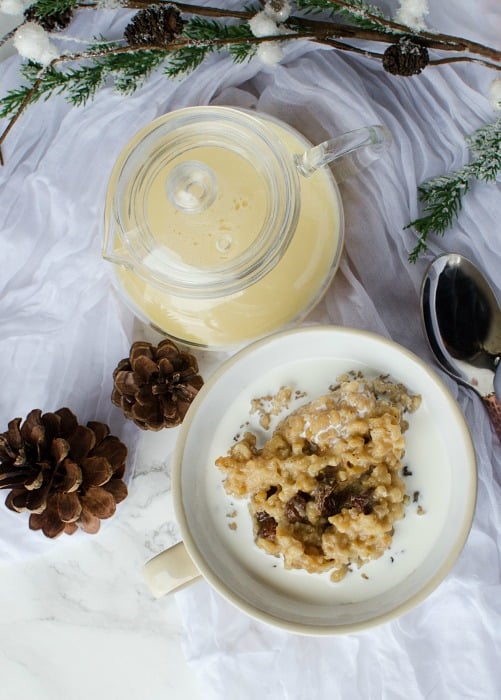 Crock Pot Eggnog Oatmeal in a white bowl next to a spoon, pinecones, and a pitcher of eggnog