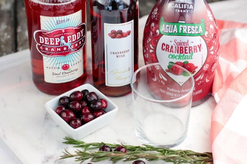 cranberry vodka, cranberry wine, and cranberry cocktail juice next to a bowl of cranberries and a glass. 