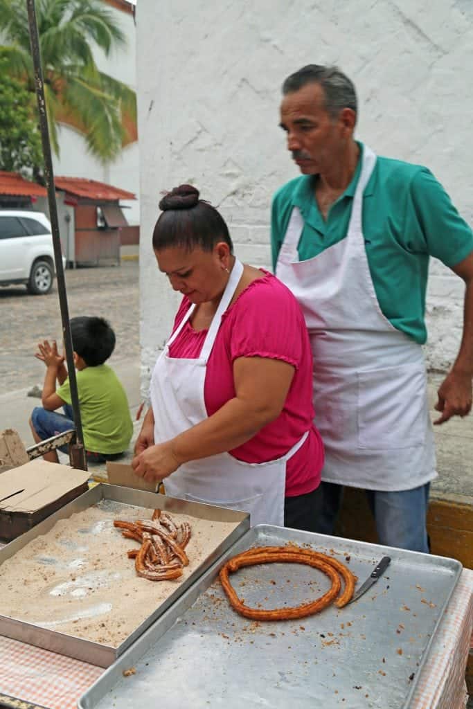 female and male next to fresh churros on a silver platter, palm trees in the background. 