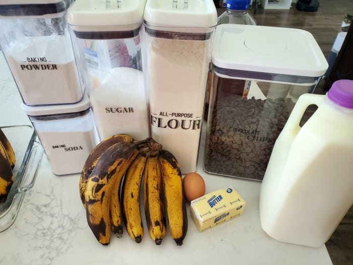Baking powder, baking soda, sugar, flour, chocolate chips, bananas, an egg, butter, and milk on a counter. 