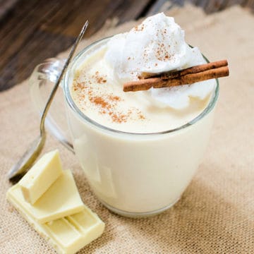 white hot chocolate in a glass coffee mug with a silver spoon, white chocolate pieces, and cinnamon sticks