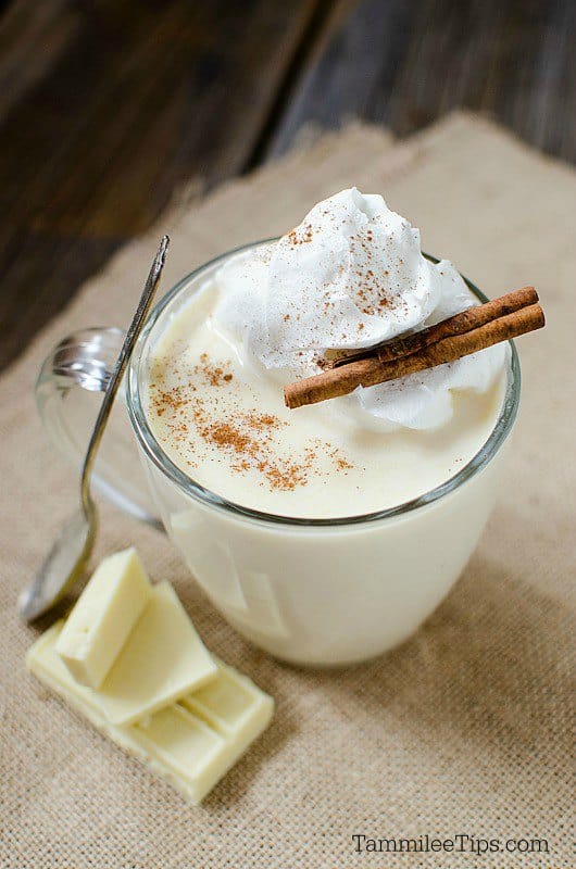 Whipped cream and cinnamon sticks garnishing a white hot chocolate in a glass mug next to a spoon