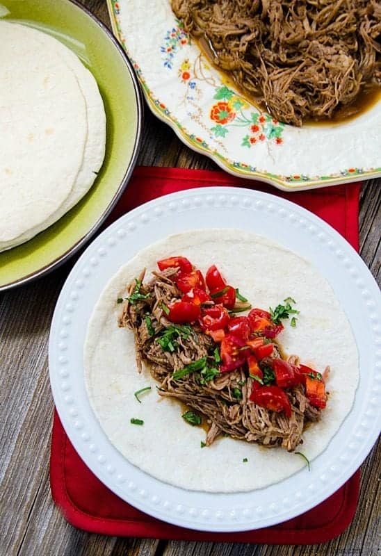 Mexican Pulled Pork, tomatoes, onions, on a tortilla on a white plate next to a serving dish of mexican pulled pork