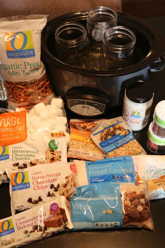 pretzels, marshmallows, chocolate chips, coconut, nuts, coconut oil, sprinkles in front of a slow cooker with mason jars in it. 