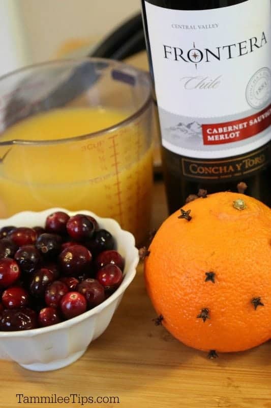 orange juice in a measuring cup, Frontera merlot bottle, bowl of cranberries, orange with cloves stuck in it. 