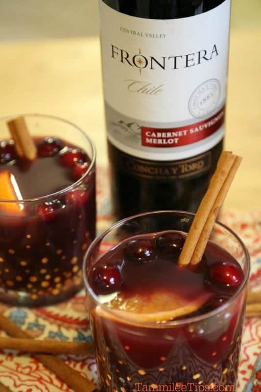 Frontera merlot wine bottle next to two glasses with red wine, cranberries, oranges, and cinnamon sticks. 