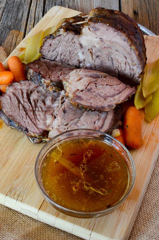 slices crockpot roast on a cutting board with carrots next to a glass bowl of gravy