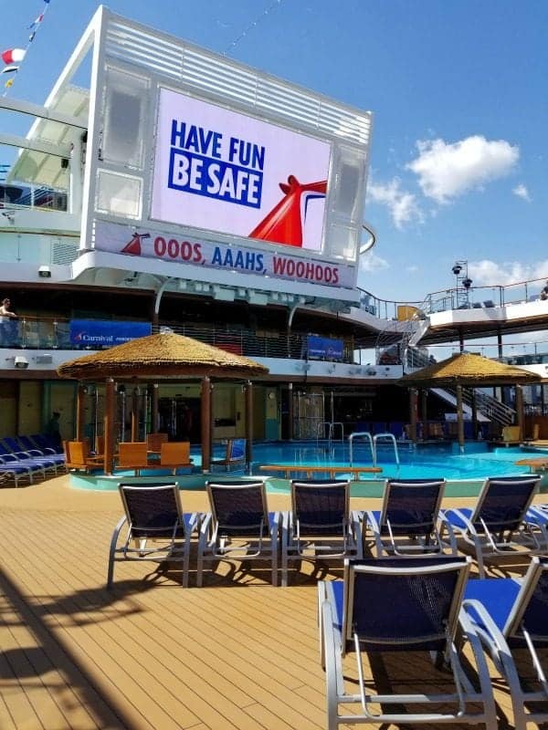 Have Fun Be Safe Carnival Cruise sign on a jumbo tv screen above the pool and lounge chairs
