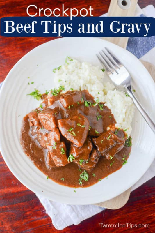 Crockpot beef tips covered in gravy next to potatoes on a white plate with a silver fork on a wood table 