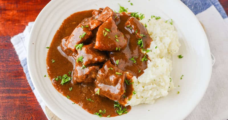 Beef tips covered in gravy next to mashed potatoes on a white plate
