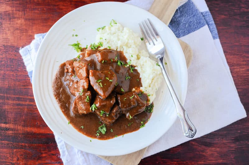 Crockpot beef tips covered in gravy with mashed potatoes on a white plate with a silver fork and cloth napkin