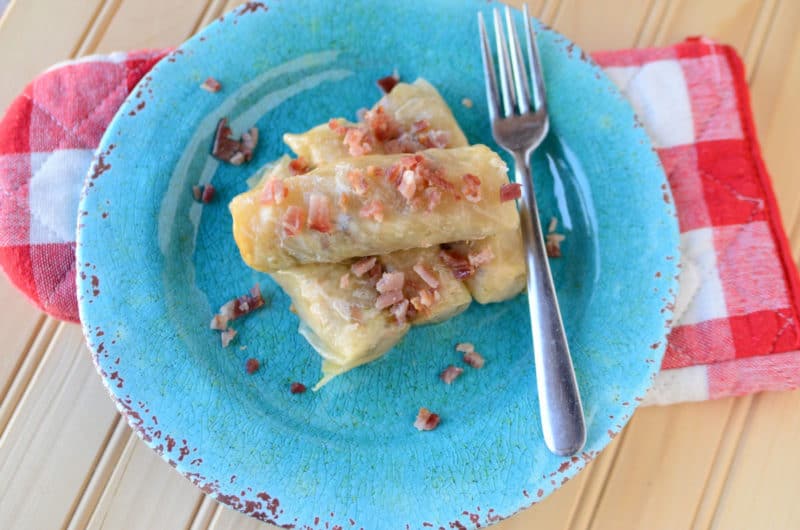 Cabbage rolls garnished with bacon on a blue plate next to a silver fork and checkered oven mit