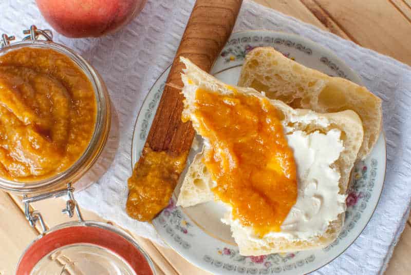  a white napkin with a wooden knife, muffin covered in peach butter, a fresh peach, and glass lidded container with peach butter in it. 