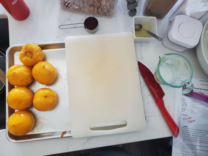 peaches on a baking sheet with a cutting board and red knife next to a measuring cup