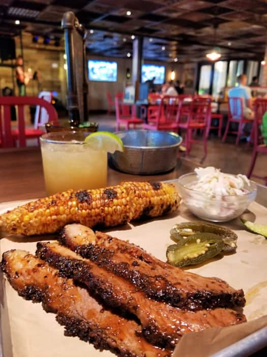 strips of beef brisket, corn, jalapenos, and a cocktail in a restaurant with red chairs and TVs on the wall