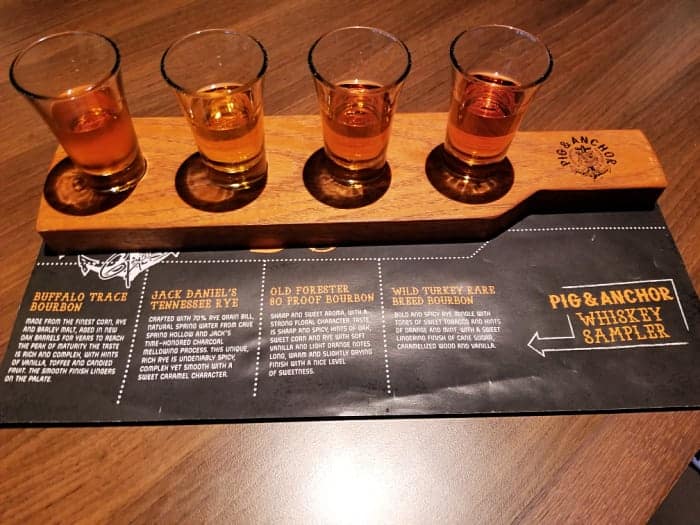 Pig & Anchor Whiskey sampler sheet below a wood board with 4 shot glasses with whiskey in them. 
