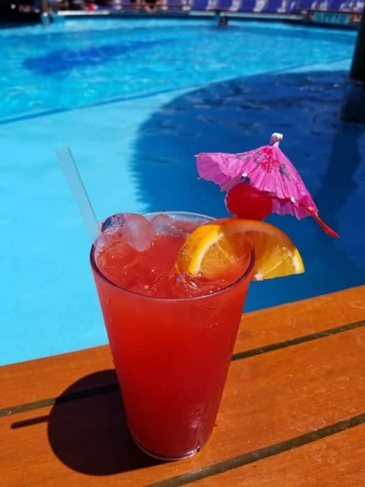 Red drink with orange wedge, cherry, and umbrella garnish next to a pool 