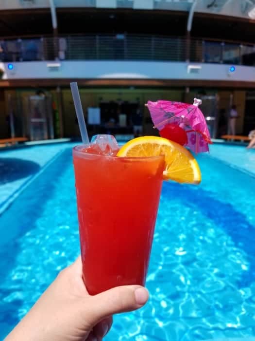 Hand holding a red drink garnished with a orange, cherry, and tropical umbrella next to a pool 