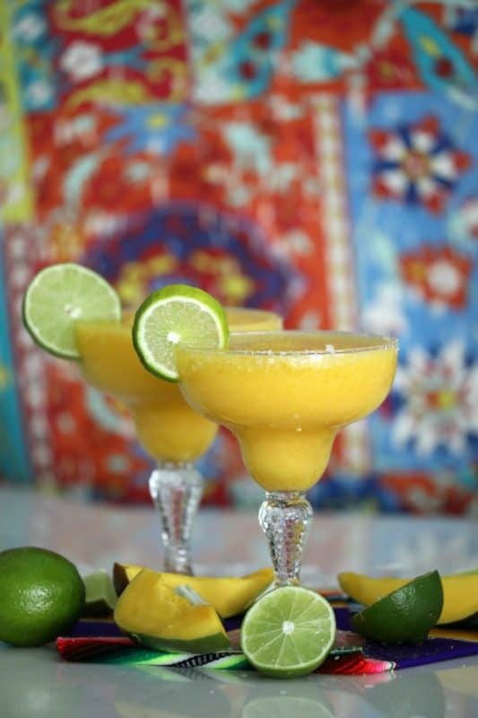 yellow margaritas in margarita glasses with lime wheels and mangos