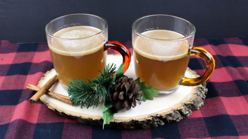 Two mugs of hot buttered rum on a wooden board with cinnamon sticks and a checkered tablecloth