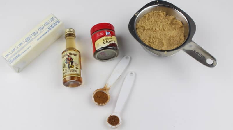 A stick of butter, spiced rum bottle, ground cloves, brown sugar, and more spices on a white counter